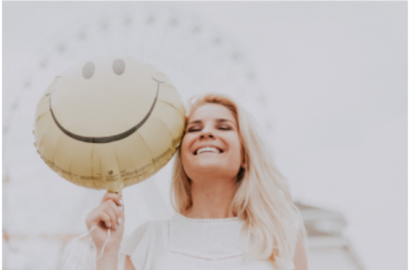 eine Frau mit blonden langen Haaren hält vor einem Riesenrad einen Smileyluftballon neben ihr Gesicht