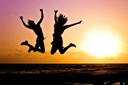 zwei Frauen springen vor Freude am Strand, im Hintergrund ein Sonnenaufgang